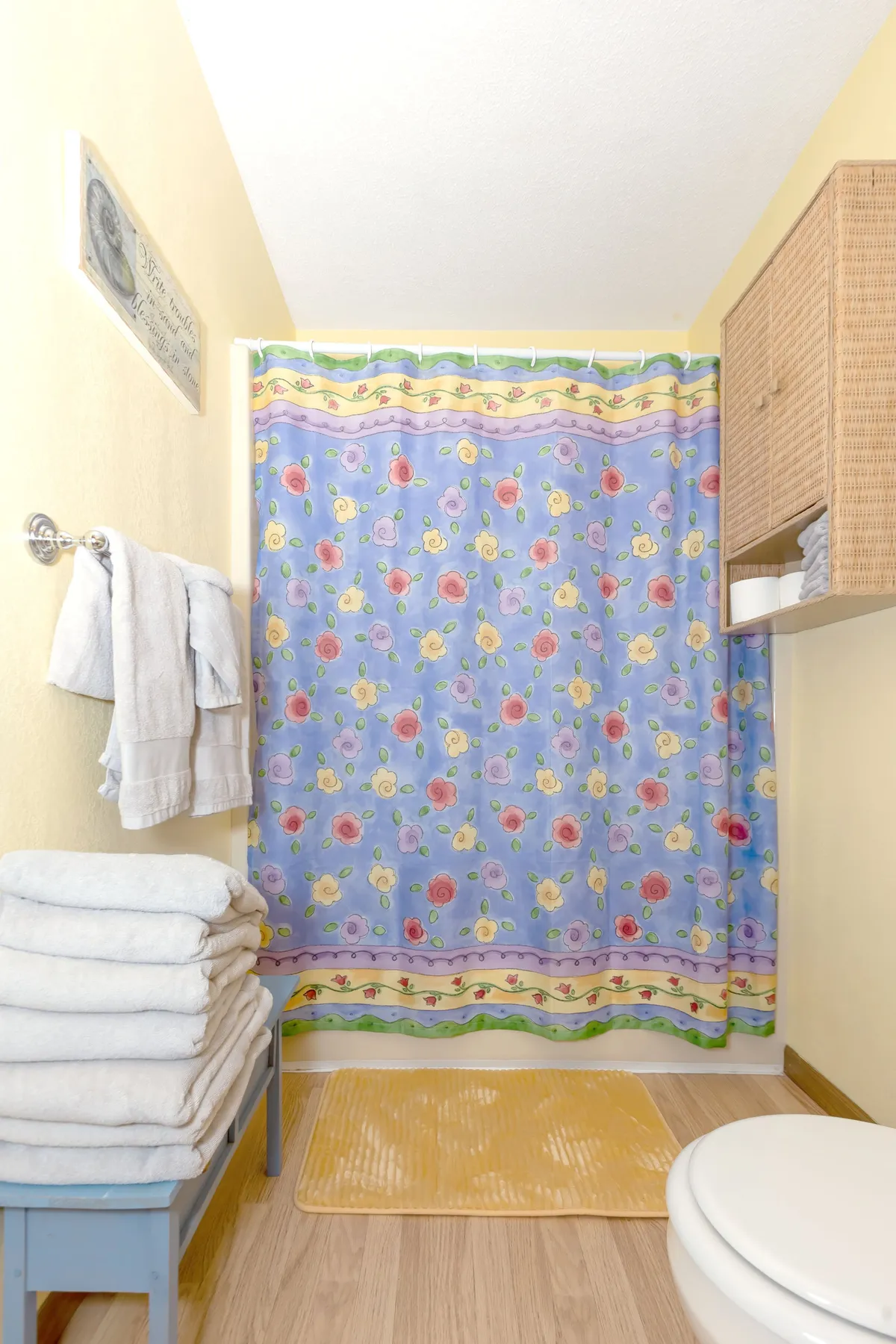 A cozy bathroom featuring a floral shower curtain, a stack of towels, a soft yellow bath mat, and a wicker storage unit. The mood is inviting and clean, ideal for self-care and relaxation.