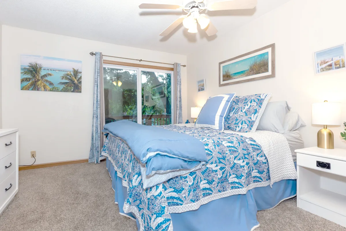 A serene bedroom featuring a bed with blue and white bedding, a large window that floods the room with natural light, and calming wall art. The view outside the window suggests a connection to the tranquil outdoors, reinforcing the peaceful atmosphere.