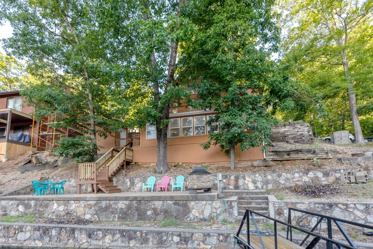 A spacious wooden deck outfitted with colorful chairs, surrounded by large trees and a stone pathway. The area is set up for gatherings, barbecues, and outdoor relaxation, with ample room for groups to spread out and enjoy the natural setting.