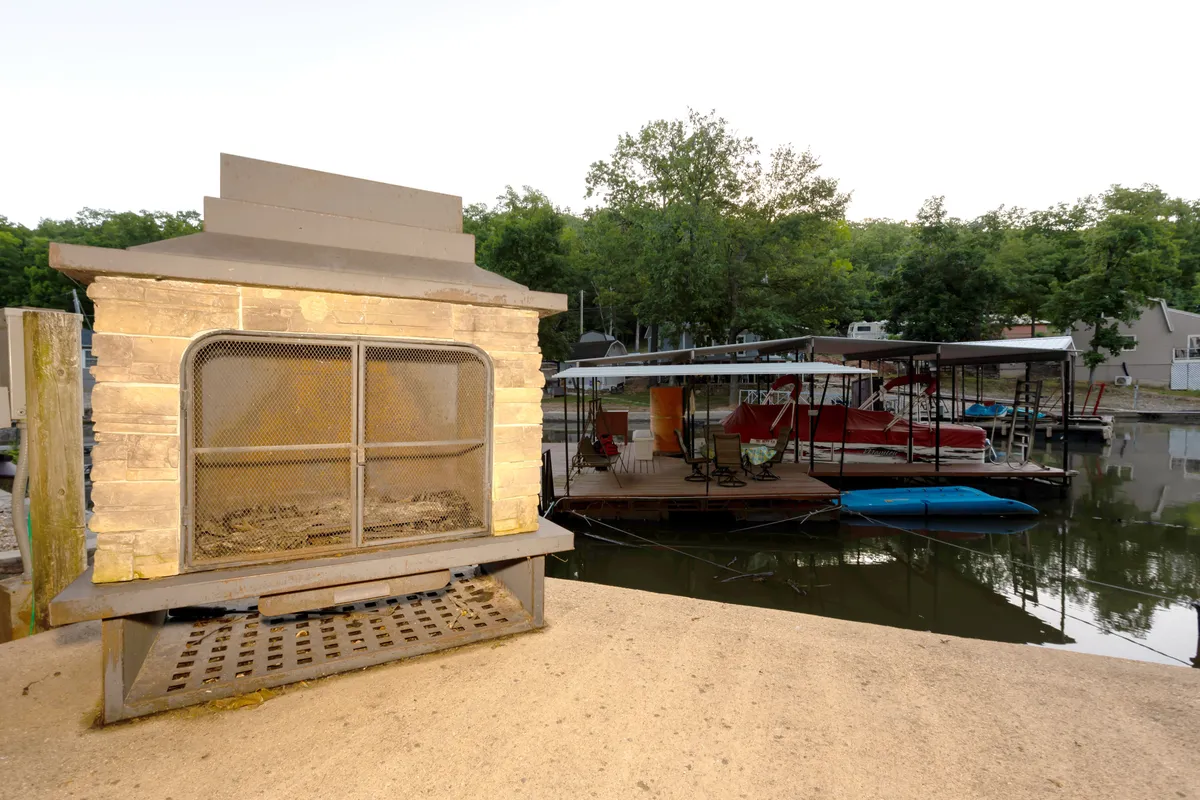 This image presents a dock with boats, a stone fireplace nearby, and a wide view of the water, emphasizing both active and relaxing lakeside experiences. The setting captures the potential for boating, fishing, evening gatherings around the fireplace, and enjoying panoramic views.