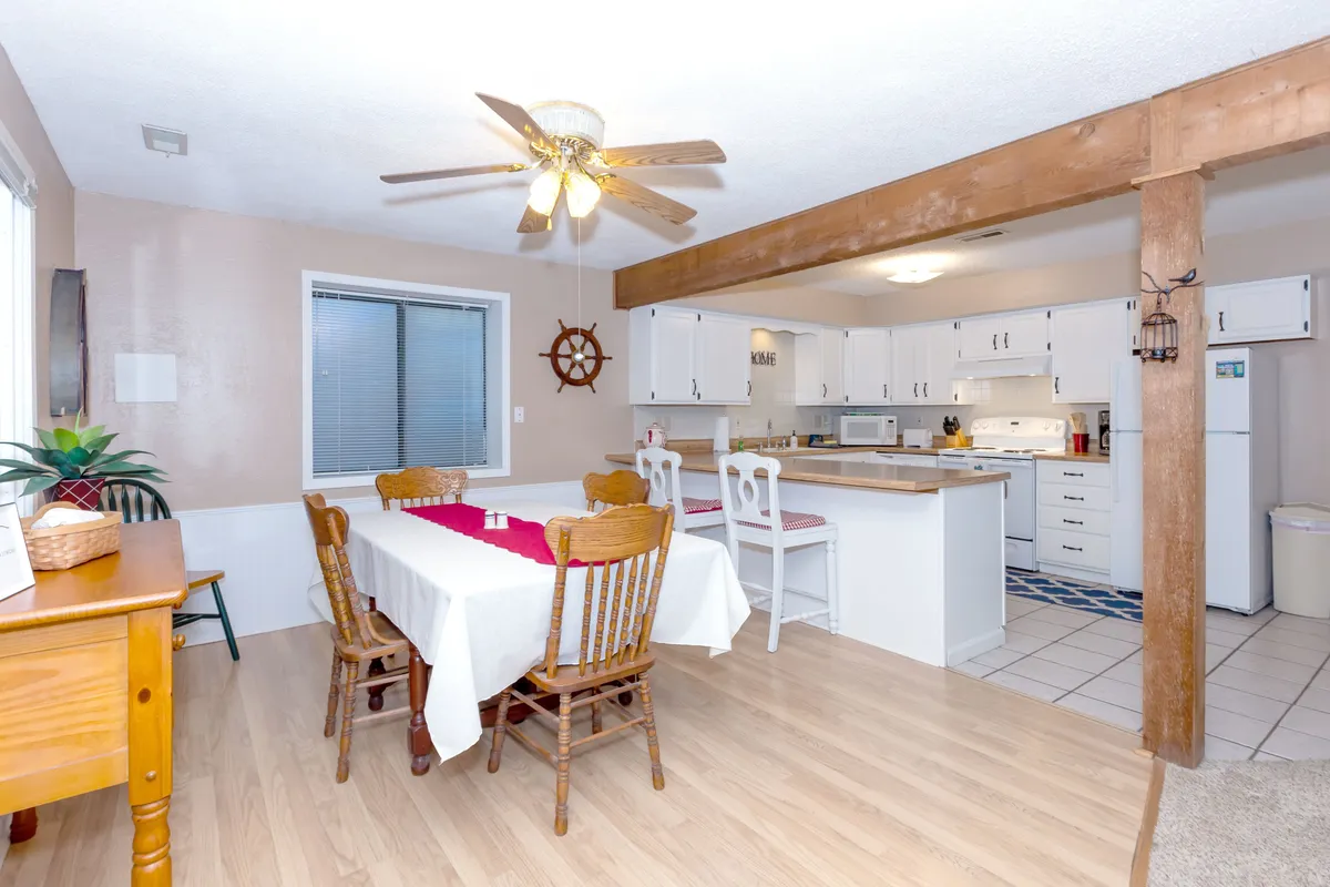 The shot centers on a dining table with a vibrant red runner, ceiling fan and wooden beam overhead, with the kitchen area visible. The inviting setup suggests occasions for family meals, celebrations, and friendly gatherings, anchoring the dining experience at the heart of the home.