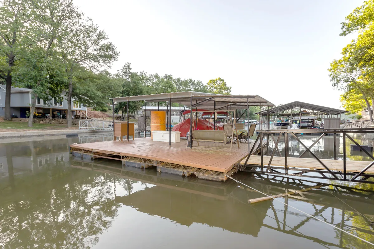 A peaceful lakeside scene centered on a wooden dock with a metal canopy, overlooking glassy water and surrounded by trees and boathouses. The dock is spacious and inviting, perfect for swimming, paddleboarding, or simply soaking up the lakeside ambiance.