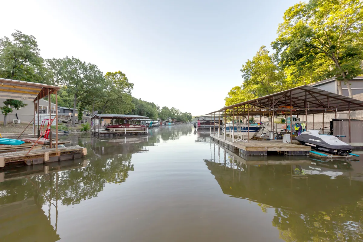 A peaceful lakeside scene with boathouses lining the shore, calm water reflecting the trees, and the soft light of either early morning or evening. The shoreline is natural and minimally developed, suggesting tranquility and opportunities for boating, fishing, and wildlife observation in a quiet cove setting.