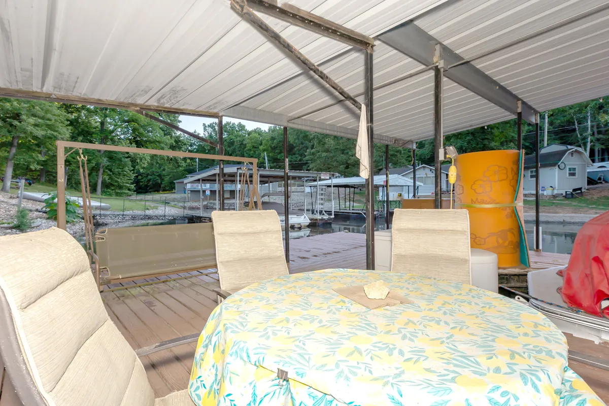 This image features a round table with a floral tablecloth and two chairs set on a patio overlooking the water. In the background, a swing and docks with boats are visible, helping to establish the relaxing lakeside setting. The scene suggests opportunities for casual dining, fishing, and enjoying the peaceful water views.